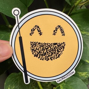 May include: A yellow smiley face sticker with a black dotted smile and eyes. The sticker is on a white background with a black drumstick.