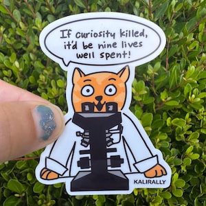 May include: A die-cut sticker featuring an orange cat wearing a white lab coat and looking through a black microscope. A speech bubble above the cat contains the text: If curiosity killed, it'd be nine lives well spent! The word KALIRALLY is printed at the bottom.