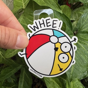 May include: A white beach ball with red, yellow, and blue sections. The ball has a cartoon face with two large eyes and the word "WHEE!" above it. The ball is on a green leafy background.