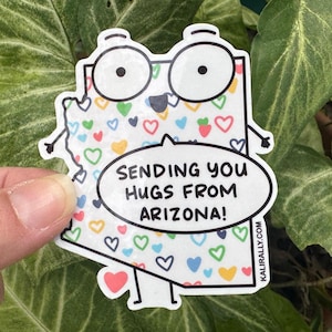 May include: A white sticker in the shape of Arizona with a cartoon face and arms, wearing glasses. The sticker is covered in colorful hearts and has a speech bubble that says "Sending you hugs from Arizona!"