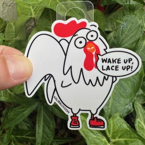 May include: White rooster sticker with red comb and wattle, wearing red shoes. The rooster is saying "Wake up, Lace up!" in a speech bubble. The sticker is on a green leafy background.