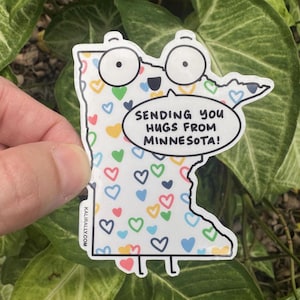 May include: A sticker in the shape of Minnesota with cartoon eyes and glasses, covered in colorful hearts. The sticker has the text "Sending you hugs from Minnesota!" in a speech bubble.