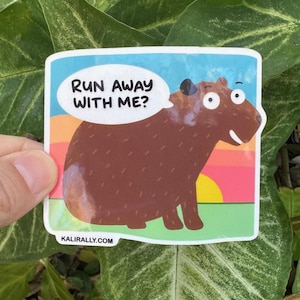 May include: A sticker featuring a cartoon capybara with a speech bubble that says "RUN AWAY WITH ME?". The capybara is brown with a white belly, set against a colorful sunset background. The website address is at the bottom.