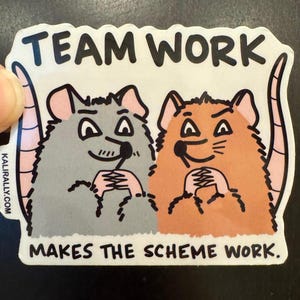 May include: A sticker with the text "TEAM WORK" above two cartoon rats holding hands. The text "MAKES THE SCHEME WORK" is below the rats. The rats are gray and orange with pink ears and tails.