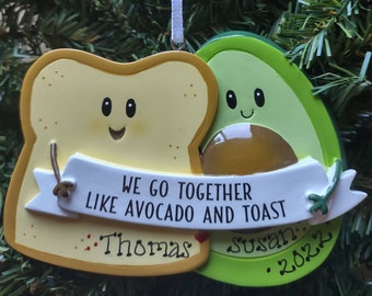 May include: A whimsical Christmas ornament featuring a slice of toast and an avocado, each with smiling faces. A banner reads "WE GO TOGETHER LIKE AVOCADO AND TOAST." Personalized with names "Thomas" and "Susan" and the year "2022".