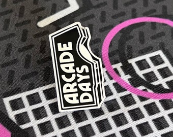 May include: Black and white enamel pin with the text "ARCADE DAYS" in a white box with a jagged edge. The pin is on a black and white patterned background with a pink circle and a blue, pink, and yellow geometric shape.