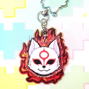 May include: A clear acrylic keyring featuring a white fox mask with red markings and a fiery red and orange outline. The keyring has a silver-coloured clasp and a flower-shaped charm.