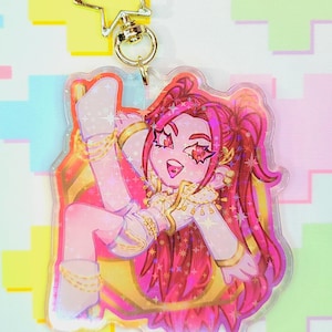May include: A clear acrylic charm featuring a cartoon character with pink hair and star-shaped eyes. The character is wearing a white outfit with gold accents and is posed with one leg raised. The charm has a gold star-shaped clasp.