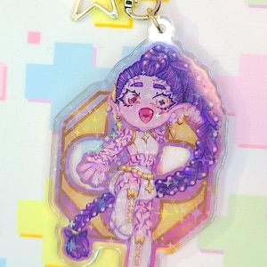 May include: A clear acrylic keychain featuring a cartoon character with purple hair and a matching braid. The character is wearing a white top and has a pink mouth. The keychain has a gold star-shaped charm and a gold clasp.