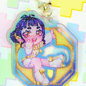 May include: A clear acrylic charm featuring a cartoon character with purple hair, freckles, and a white and gold outfit. The charm has a gold star-shaped clasp and a gold ring. The background is a colorful pixelated design.