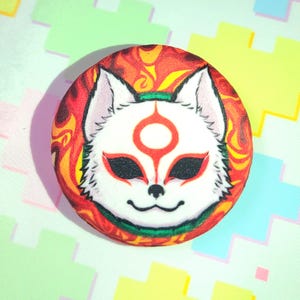 May include: A round button featuring a white fox mask design with red accents and a fiery orange and yellow background. The fox has red markings around the eyes and forehead, with a green band across the forehead. The button is a vibrant accessory.