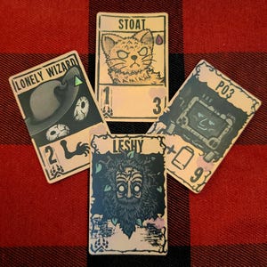 May include: Four illustrated game cards with unique designs and text. The cards feature illustrations of a cat, a wizard, a creature, and a machine. Each card has a different name: "STOAT", "LONELY WIZARD", "LESHY", and "P03". The cards are arranged on a red and black checkered surface.