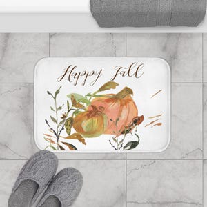 Op de afbeelding: Een rechthoekige badmat met een witte achtergrond voorzien van een aquarelillustratie van oranje en groene pompoenen omringd door herfstbladeren. De tekst "Happy Fall" staat in een bruin cursief lettertype boven het ontwerp.