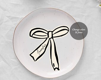 May include: A white plate with a black and white illustration of a bow with a ribbon that says "Jennifer".