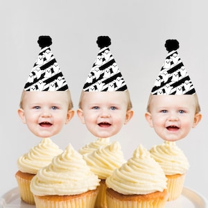 May include: Three cupcakes with white frosting topped with ghost-themed party hats. Each hat has a black and white striped pattern with white ghosts. The hats have a black pom-pom on top. Each cupcake has a baby's face on top of the hat.