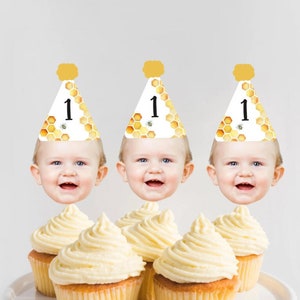 Puede incluir: Tres cupcakes con glaseado blanco cubiertos con la cara de un bebé que lleva un gorro de fiesta de panal amarillo y blanco con el número "1" en él.