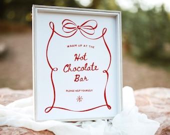 Può includere: Un cartello bianco incorniciato con scritte rosse e un bordo decorativo. Il cartello recita "Warm up at the Hot Chocolate Bar. Please help yourself!" Sono presenti anche un fiocco rosso e un fiocco di neve. Il cartello è appoggiato su una superficie di pietra con un panno bianco.