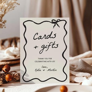 K&ouml;nnte beinhalten: Ein rechteckiges Schild mit schwarzem, wellenf&ouml;rmigem Rand und einer Schleife oben. Auf dem Schild steht "Cards + gifts" und "THANK YOU FOR CELEBRATING WITH US!" mit den Namen "Katie + Martin" unten. Das Schild steht auf einem Tisch mit Herbstdekorationen.