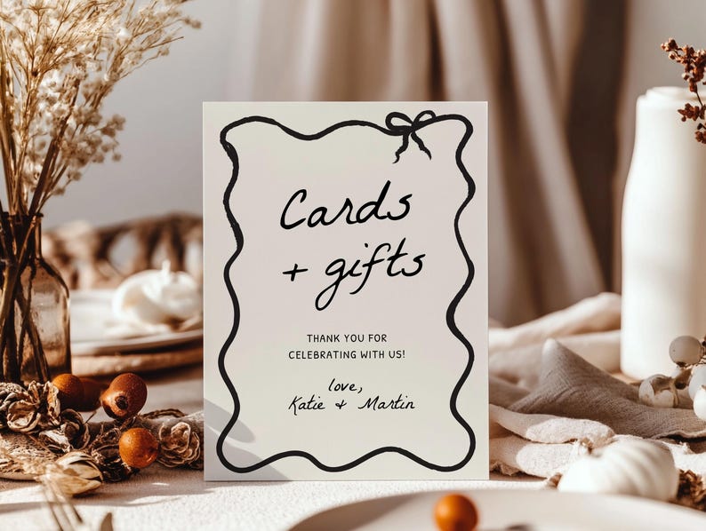 K&ouml;nnte beinhalten: Ein rechteckiges Schild mit schwarzem, wellenf&ouml;rmigem Rand und einer Schleife oben. Auf dem Schild steht "Cards + gifts" und "THANK YOU FOR CELEBRATING WITH US!" mit den Namen "Katie + Martin" unten. Das Schild steht auf einem Tisch mit Herbstdekorationen.