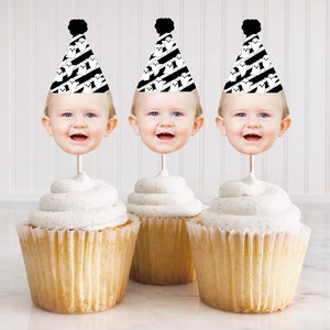 May include: Three cupcakes with white frosting topped with ghost-themed party hats with a baby's face in each hat. The hats are black and white with a black pom-pom on top.