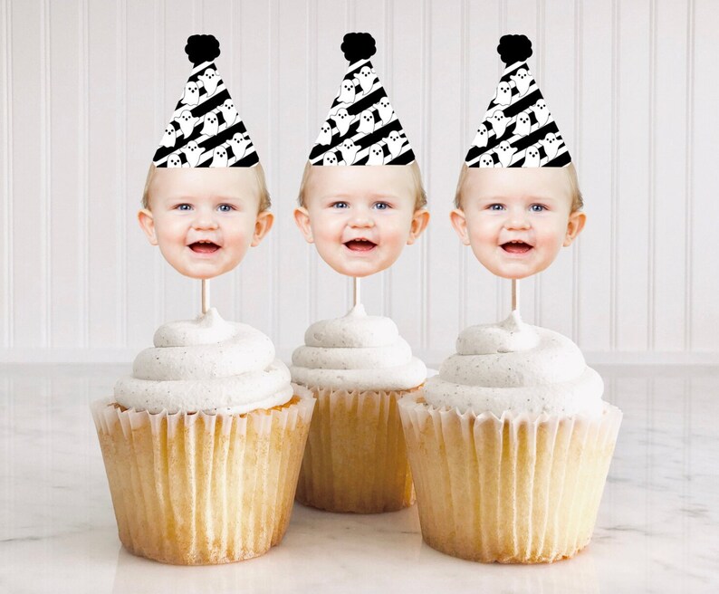 May include: Three cupcakes with white frosting topped with ghost-themed party hats with a baby's face in each hat. The hats are black and white with a black pom-pom on top.