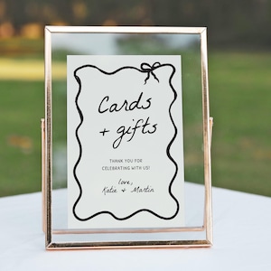 K&ouml;nnte beinhalten: Ein klarer Glas- und goldfarbener Metallrahmen mit den Worten "Cards + gifts" in schwarzer Schrift. Das Schild enth&auml;lt den Text "Thank you for celebrating with us!" und die Namen "Katie + Martin".