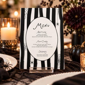 May include: A rectangular menu card featuring a black and white striped background with spider web and spider illustrations. The center contains an oval white space with the text Menu, Appetizers, Main Course, and Desserts listed in black.