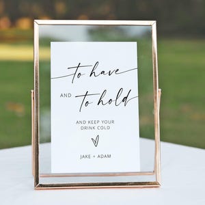 May include: A framed sign with a clear glass front and a gold-colored metal frame. The white card inside has elegant black script that reads "to have and to hold" and "and keep your drink cold." The names "Jake + Adam" are at the bottom.