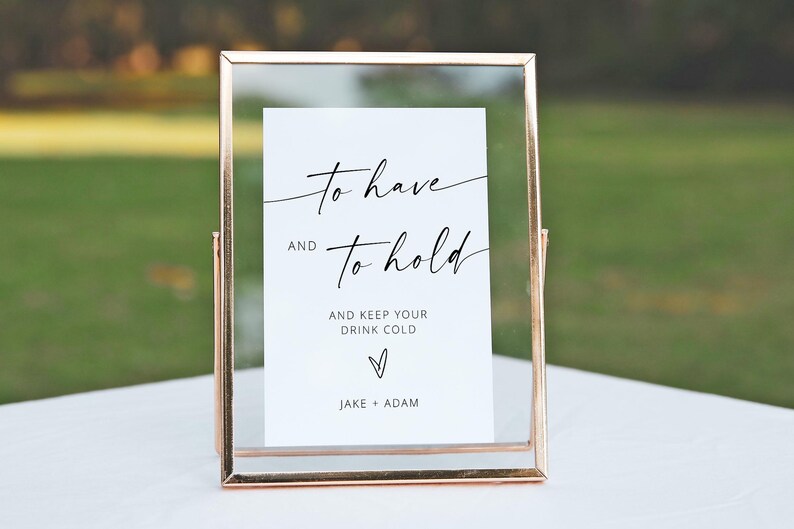 May include: A framed sign with a clear glass front and a gold-colored metal frame. The white card inside has elegant black script that reads "to have and to hold" and "and keep your drink cold." The names "Jake + Adam" are at the bottom.