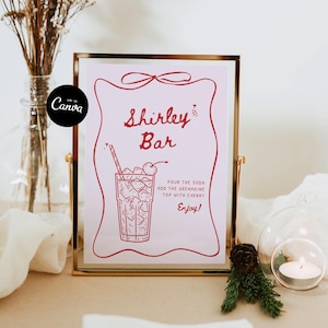 May include: A framed sign with the words "Shirley Bar" in red script. The sign features a drawing of a cocktail with ice and a cherry, and instructions for the drink. The frame is gold-colored.