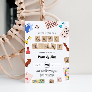 May include: A white invitation card with colorful illustrations of board games, such as chess, checkers, and dice. The text reads "You're invited to a Game Night housewarming for Pam & Jim, Saturday, May 23rd, 20XX at 4:00 PM. The Reynolds Residence, 1234 Main St, Los Angeles. Kindly RSVP to Emma (345) 567.890.7654."