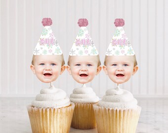 May include: Three cupcakes with white frosting topped with a baby's face wearing a pink and white snowflake party hat with the word "one" on it.