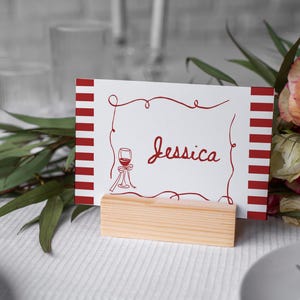May include: A white place card with red stripes and the name "Jessica" in cursive script. A red wine glass illustration is on the card. The card is held in a small, natural wood block. A bouquet of roses and greenery is in the background.
