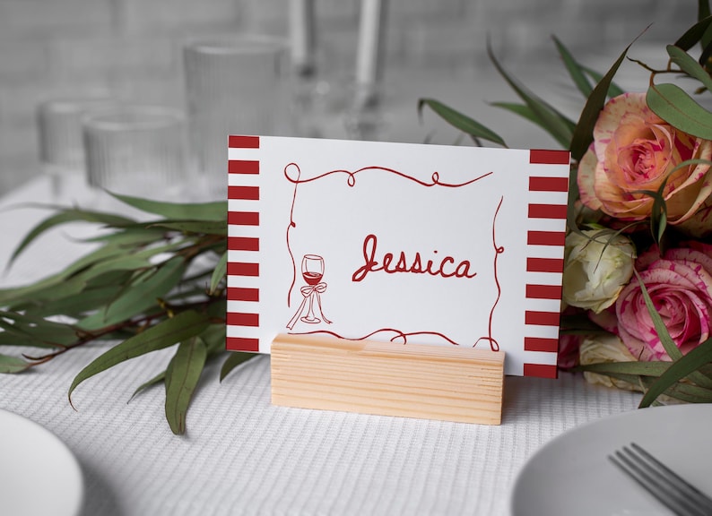May include: A white place card with red stripes and the name "Jessica" in cursive script. A red wine glass illustration is on the card. The card is held in a small, natural wood block. A bouquet of roses and greenery is in the background.