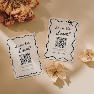 May include: Two white cards with black wavy borders and the text "Share the Love" with a QR code and the text "Scan the code to share your photos with us!" and the names "xoxo, Lia + Mike". Dried flowers are scattered around the cards.