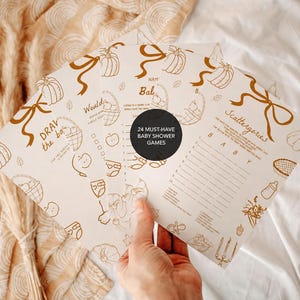 May include: A set of off-white paper baby shower game cards featuring brown line art illustrations of pumpkins, ribbons, and baby items. The text includes titles like Draw the Baby and Scattergories, with a black circular overlay stating 24 MUST-HAVE BABY SHOWER GAMES.
