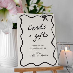 K&ouml;nnte beinhalten: Eine wei&szlig;e Karte auf einer kleinen Holzstaffelei mit schwarzem handgeschriebenem Text, der "Cards + gifts" lautet. Die Karte hat einen dekorativen schwarzen Rand und eine Schleife oben. Der Text "Thank you for celebrating with us! love, Katie & Martin" steht ebenfalls auf der Karte.