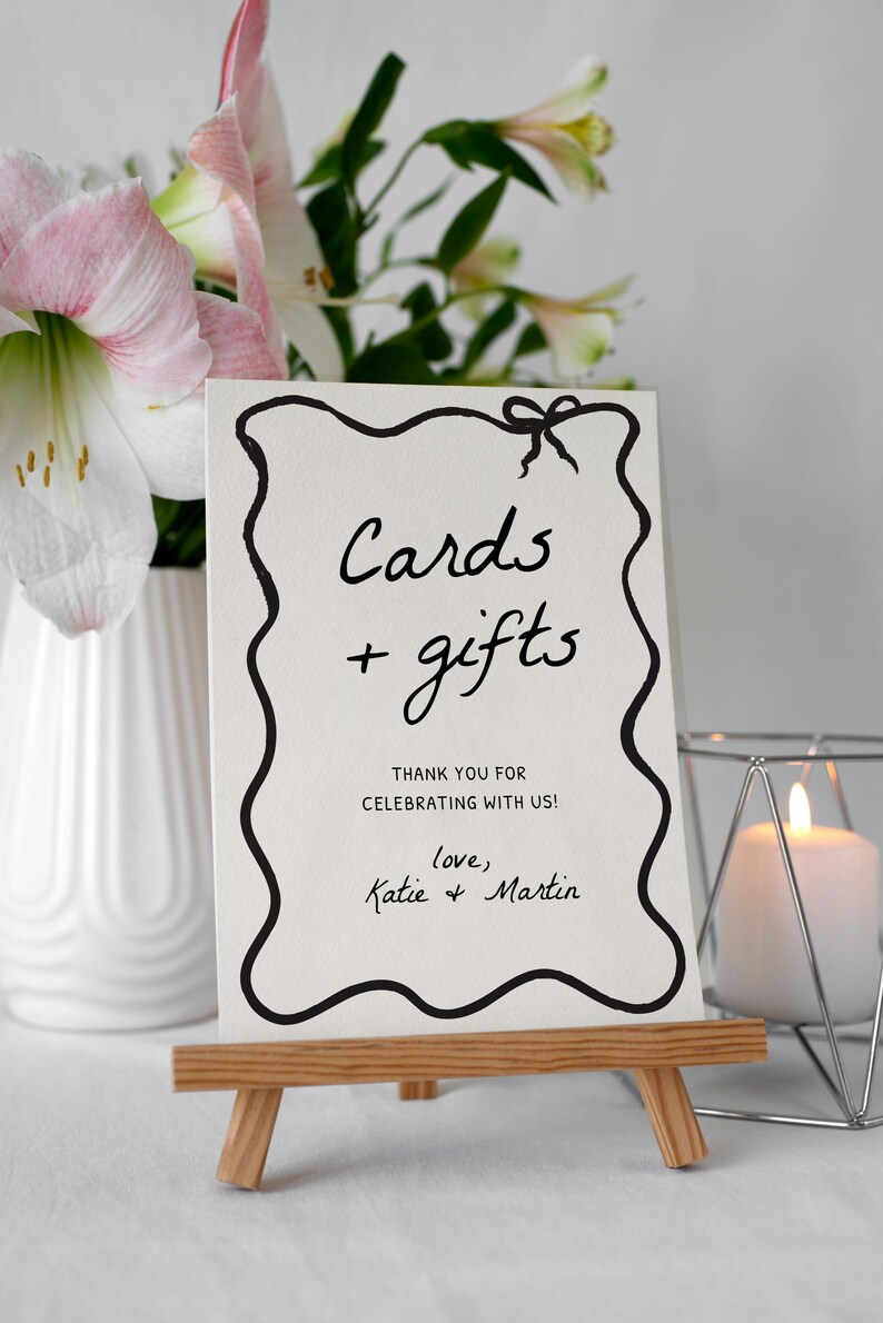 K&ouml;nnte beinhalten: Eine wei&szlig;e Karte auf einer kleinen Holzstaffelei mit schwarzem handgeschriebenem Text, der "Cards + gifts" lautet. Die Karte hat einen dekorativen schwarzen Rand und eine Schleife oben. Der Text "Thank you for celebrating with us! love, Katie & Martin" steht ebenfalls auf der Karte.