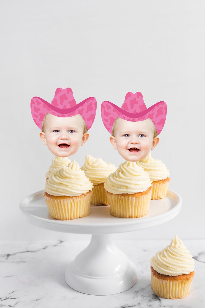 May include: Two cupcakes with white frosting topped with pink cowboy hats with a cow print design. The hats have a baby's face peeking out from underneath. A third cupcake with white frosting is on a white cake stand.