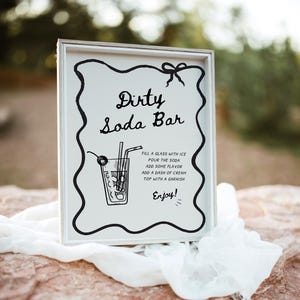 Op de afbeelding: Een ingelijst bord met de woorden "Dirty Soda Bar" in elegant schrift. Het bord bevat instructies voor het maken van een frisdrank, een tekening van een frisdrank en een decoratieve zwarte rand. Het bord rust op een witte doek.