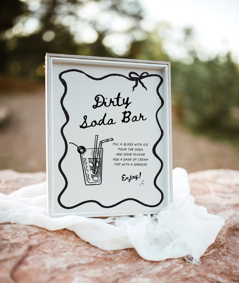 Op de afbeelding: Een ingelijst bord met de woorden "Dirty Soda Bar" in elegant schrift. Het bord bevat instructies voor het maken van een frisdrank, een tekening van een frisdrank en een decoratieve zwarte rand. Het bord rust op een witte doek.