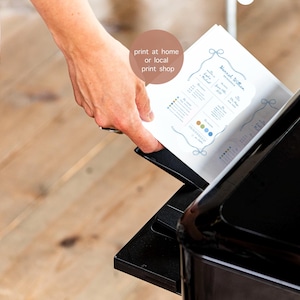 May include: A white rectangular paper document with blue text and a decorative border being fed into a black printer. The document features the text 'print at home or local print shop' in a circular brown speech bubble overlaying the paper.