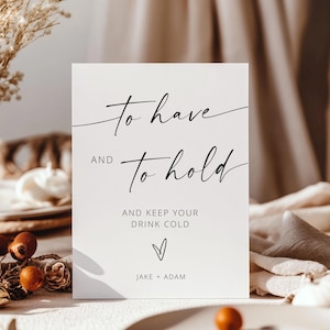 May include: A white rectangular sign with black script that reads "To have and to hold" and "And keep your drink cold." A heart and names "Jake + Adam" are below the text. The sign is on a table with decorative elements.