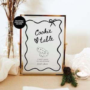 May include: A gold-framed sign with the words "Cookie table" in a handwritten font, along with a heart symbol and a drawing of two cookies. The sign also includes the text "A sweet ending to a new beginning" and the names "Sarah + Jason."