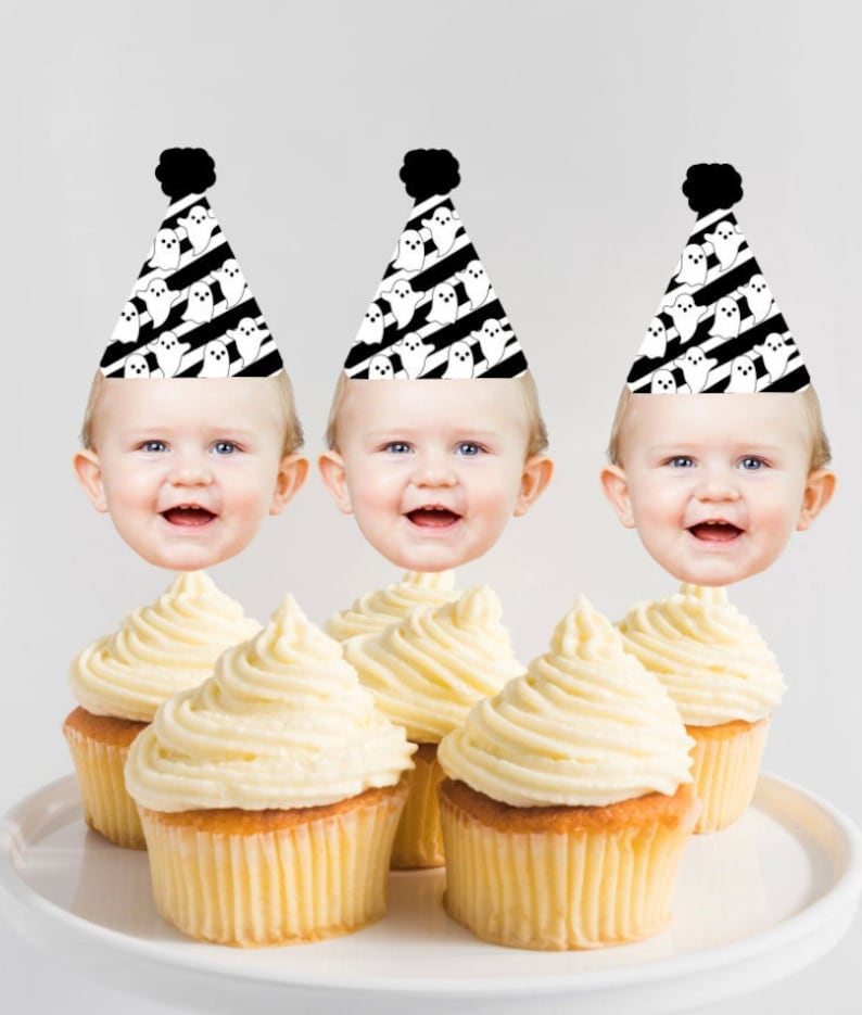 May include: Three cupcakes with white frosting topped with ghost-themed party hats. Each hat has a black and white striped pattern with white ghosts. The hats have a black pom-pom on top. Each cupcake has a baby's face on top of the hat.