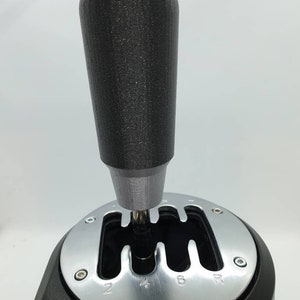 May include: A black gear knob with a textured surface, attached to a black and silver gear shifter with a 6-speed pattern and a reverse gear.