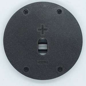 May include: A circular black plastic plate featuring a textured surface, four mounting holes near the perimeter, a central vertical slot with a visible black cord, and embossed plus and minus symbols positioned above and below the central opening.