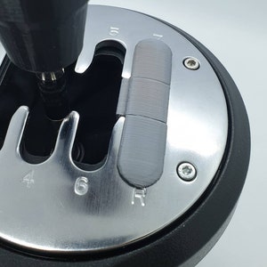 May include: A silver gear shift knob with black trim and a gray plastic insert. The knob has numbers 2, 4, 6, and R printed on it. The insert has the numbers 5 and 7 printed on it. A drill is being used to install the insert.