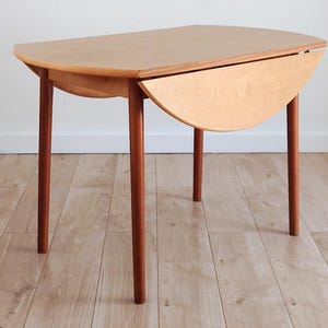 May include: A light brown wooden drop-leaf table with a smooth, oval-shaped top and four cylindrical legs. The table features one side leaf folded down, revealing the grain of the wood and the structural support mechanism underneath the surface.