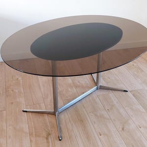 May include: An oval dining table featuring a translucent brown glass top with a solid black oval centre inlay. The table is supported by a silver-toned metal frame consisting of two vertical pillars connected by a horizontal bar and four legs.